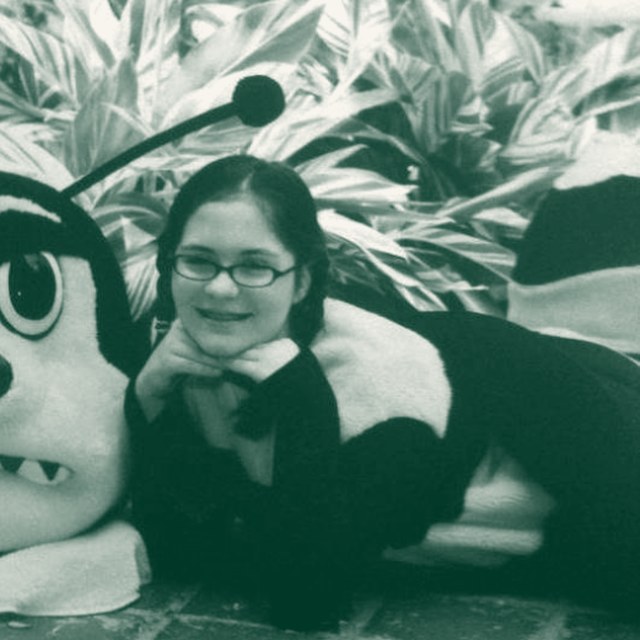 Kellie as a kid in glasses and braids, posing with a bee mascot