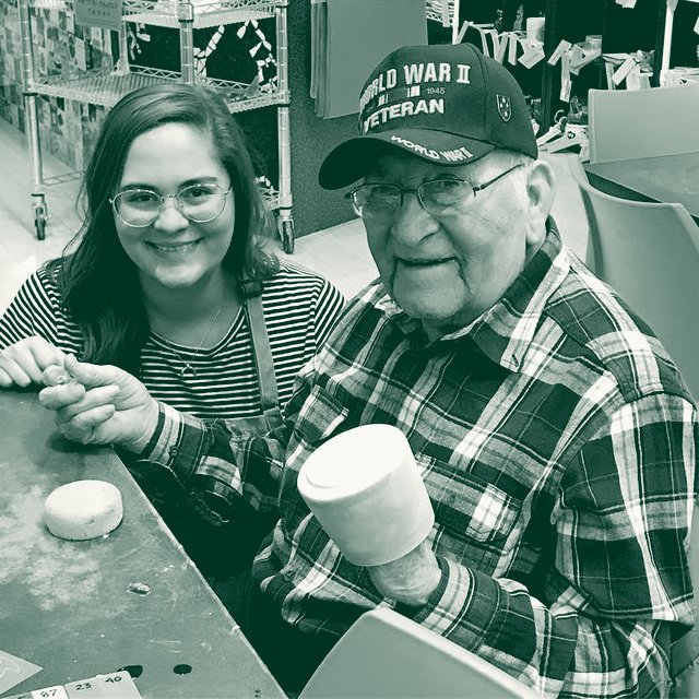 Kellie smiling beside an elderly veteran as they paint ceramics together at a craft table